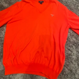 Orange tröja från GANT - Säljer en snygg orange tröja från GANT med v-ringning och broderad logga på bröstet. Tröjan har långa ärmar och ribbade kanter, perfekt för en stilren look.