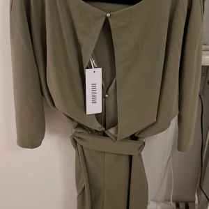 Olivgrön klänning  - Elegant olivgrön klänning med trekvartsärm och knytband i midjan. Klänningen har en stilren design med en rund halsringning och en diskret slits framtill. Perfekt för en chic och sofistikerad look.