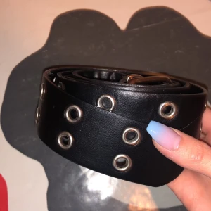 Svart bälte med nitar - Snyggt svart bälte med silverfärgade nitar. Perfekt för att ge en edgy touch till din outfit. Tillverkat i skinnliknande material med flera justeringshål för bästa passform.