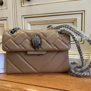 Beige Mini Kensington Bag från Kurt Geiger - Mini Kensington bag från Kurt Geiger med quiltad design och en cool kedjerem. Färg Camel. Väskan har en unik detalj på framsidan och är perfekt för att ge din outfit en stilfull touch. Den är kompakt men rymmer det viktigaste för en dag ute.