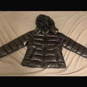 Svart dunjacka med huva - Snygg svart dunjacka med huva och dragkedja. Perfekt för kyliga dagar med sin pufferdesign som håller värmen. Jackan har långa ärmar och en glansig yta som ger en modern look.