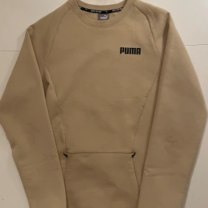 Beige tröja från Puma - Säljer en stilren beige tröja från Puma med lång ärm och rund hals. Tröjan har en diskret Puma-logga på bröstet  Perfekt för en avslappnad stil.