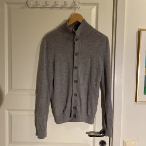 Grå cardigan Suitsupply - Säljer en stilren grå cardigan från Suitsupply storlek S i  merinoull. Koftan har en hög ribbad krage med knappar hela vägen. Trendigt och stilrent plagg som passar både med skjorta och ensamt. Plagget är u mycket bra skick utan tecken på användning.