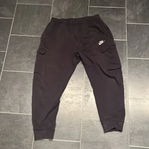 Svarta mjukisbyxor från Nike - Säljer ett par svarta mjukisbyxor från Nike med elastisk midja och praktiska cargofickor på sidorna. Perfekta för en avslappnad stil eller träning. Byxorna har en broderad Nike-logga på ena benet. Är storlek M men sitter perfekt för storlek S 