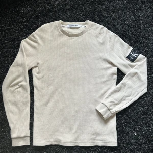 Beige långärmad tröja från Calvin Klein Jeans - Snygg beige långärmad tröja från Calvin Klein Jeans med ett diskret rutmönster. Tröjan har en rund halsringning och en svart logotypdetalj på ärmen. Perfekt för en stilren och avslappnad look.