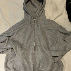 Grå hoodie från Weekday - Säljer en stilren grå hoodie från Weekday i storlek L. Den har en dragkedja framtill och en praktisk huva med snören. Perfekt för en avslappnad look.