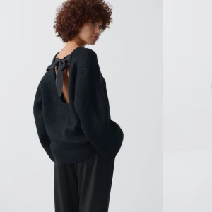Svart stickad tröja med rosett - Snygg svart stickad tröja med öppen rygg och en stor rosett som detalj. Tröjan har långa ärmar och en lös passform, perfekt för en stilren look.