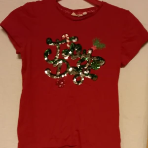 Röd t-shirt med paljetter från KappAhl - Säljer en röd t-shirt från KappAhl med glittrande paljetter som bildar texten 'Jolly Days'. Perfekt för julens festligheter. Kortärmad och bekväm passform. Storlek 146-152 