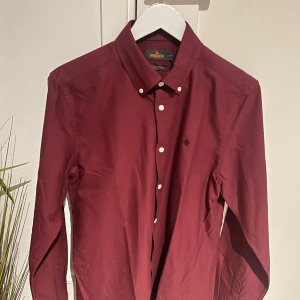 Vinröd skjorta från Morris - Snygg vinröd skjorta från Morris med klassisk krage och knappar framtill. Skjortan har en liten broderad logga på bröstet och är tillverkad i ett mjukt material.