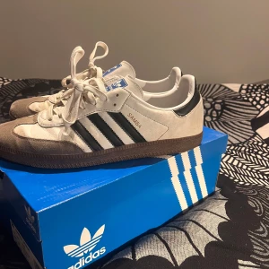 Adidas Samba sneakers - Klassiska Adidas Samba sneakers i vitt och svart med bruna sulor. Skorna har snörning och de ikoniska tre ränderna på sidan. Perfekta för en sportig och stilren look. Väl använda och därför säljer jag dem för billigt pris