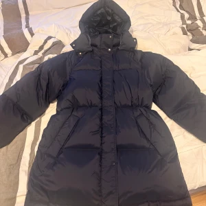  dunjacka från GANT - Säljer en stilren mörkblå dunjacka från GANT med huva och dragkedja. Perfekt för kalla dagar med sin pufferdesign som håller dig varm och bekväm. Jackan har långa ärmar och en klassisk look. Jackan är aldrig använd den är köpt på johnells 