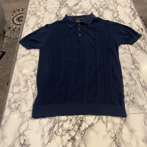 Mörkblå pikétröja från Oscar Jacobson - Snygg mörkblå pikétröja från Oscar Jacobson, tillverkad i Italien av 100% bomull. Tröjan har korta ärmar och ett klassiskt pikékrage med knappar framtill. Perfekt för en stilren look.