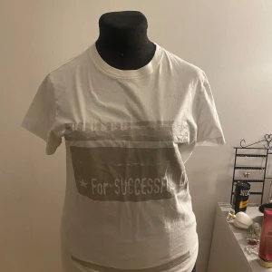 Vit t-shirt med tryck - Säljer en vit t-shirt med ett tryck i grått och texten 'For SUCCESSFUL'. T-shirten har en rund hals och korta ärmar. Perfekt för en avslappnad stil.