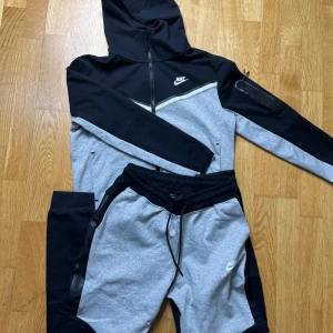 Svart och grå hoodie från Nike - Snygg och bekväm hoodie från Nike i svart och grått. Den har en dragkedja framtill och en praktisk huva. Perfekt för träning eller en avslappnad dag. Matchande byxor ingår. Storlek S men kan även passa xs.