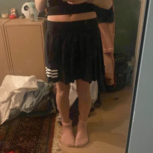 Svart kjol från Adidas - Snygg svart kjol från Adidas med vita ränder på sidan. Kjolen har en plisserad design och är tillverkad i Primegreen-material. Perfekt för en sportig look. Inbyggda shorts väldigt bekväm. Använd några gånger. 