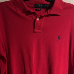 Röd pikétröja från Ralph Lauren - Säljer en klassisk röd pikétröja från Ralph Lauren med broderad logga på bröstet. Tröjan har en stilren design med krage och knappar framtill. Säljer denna då den är lite för stor för min smak. Men gillar du en Piké som inte sitter allt för tight är denna nått för dig! Kan gå ner i pris vid snabb affär