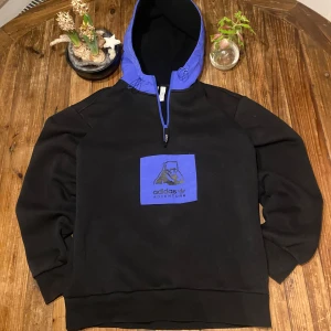 Svart och lila hoodie från Adidas Adventure - Säljer en cool svart hoodie från Adidas Adventure med en blå huva och ett stort blått tryck på framsidan. Hoodien har en justerbar dragsko i huvan och är perfekt för en avslappnad stil. Stor i storlek (passar S)