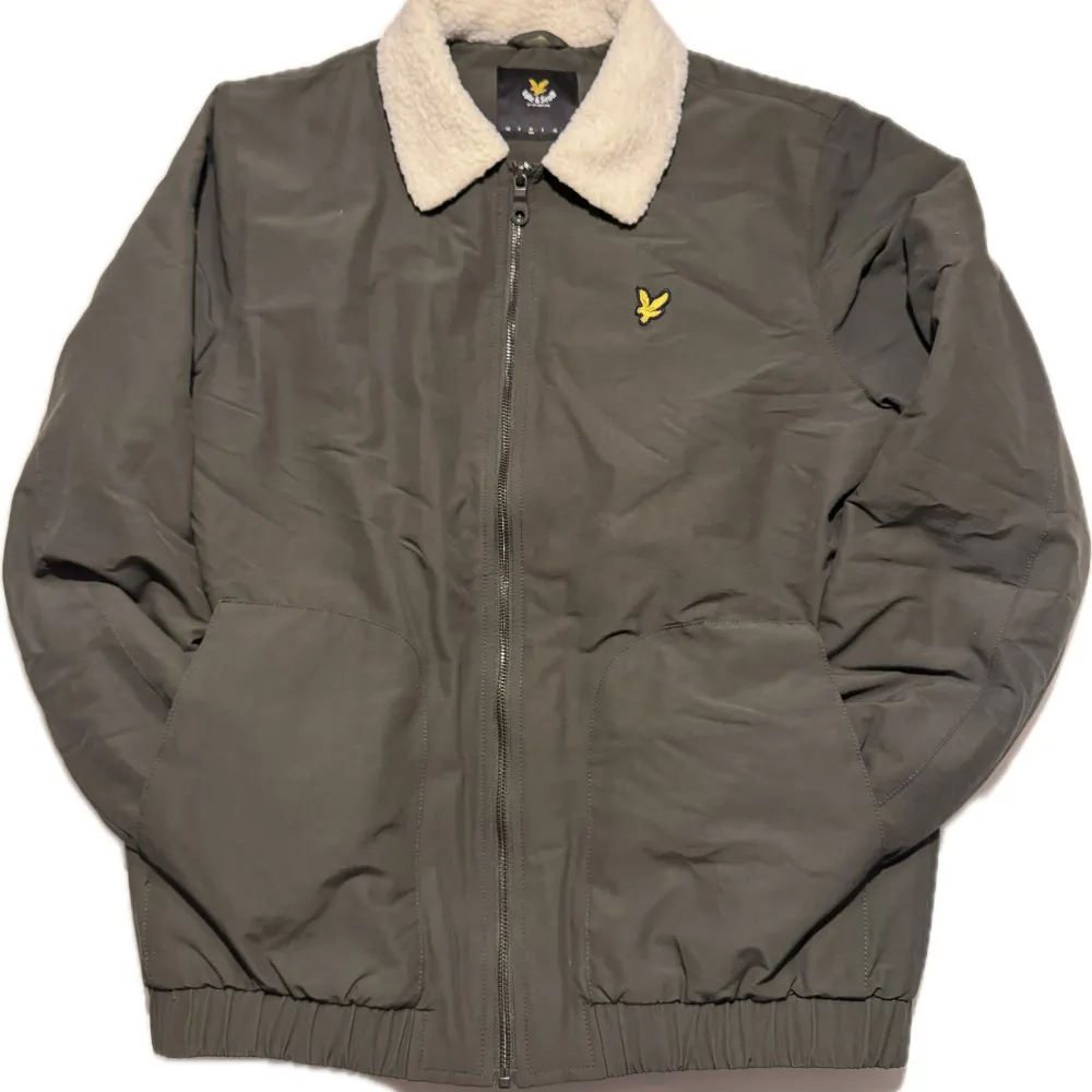 Säljer en snygg grön jacka från Lyle & Scott med mjukt foder och dragkedja. Jackan har en krage med beige foder och ett broderat gult emblem på bröstet. Perfekt för kyligare dagar!. Takit.