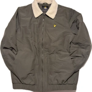 Grön jacka med foder från Lyle & Scott - Säljer en snygg grön jacka från Lyle & Scott med mjukt foder och dragkedja. Jackan har en krage med beige foder och ett broderat gult emblem på bröstet. Perfekt för kyligare dagar!