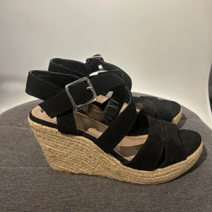 Svarta kilklackssandaler med spänne - Snygga svarta sandaler med kilklack och justerbara spännen. Sandalerna har en flätad sula i naturmaterial och korsade remmar i mocka. Perfekta för sommarens alla tillfällen. Köpta på din sko 