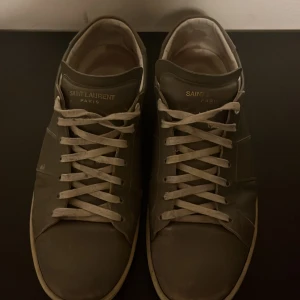 Grå sneakers från Saint Laurent - Snygga grå sneakers från Saint Laurent med klassisk snörning och stilren design. Finns små skador runt om skorna och sulan är ganska smutsig, men annars i ett 7/10 skick, sällsynt par eftersom de ej säljs längre, nypris finns då inte. Storlek 40,5 i italiensk storlek, sitter mer som 42/43.