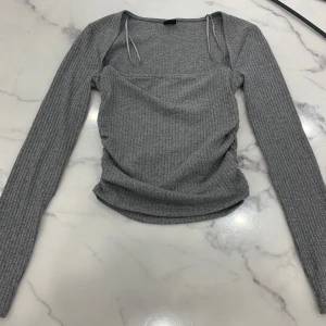 Grå ribbad tröja från Gina Tricot - Säljer en stilren grå ribbad tröja från Gina Tricot. Tröjan har långa ärmar, en snygg fyrkantig halsringning och rynkade detaljer längst sidan.