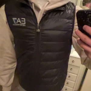 Svart dunväst från Emporio Armani - Snygg svart dunväst från Emporio Armani med EA7-logga på bröstet. Västen har en dragkedja framtill och är perfekt för lager-på-lager-stil. Den är både stilren och funktionell.