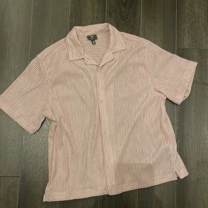 Rosa/beige kortärmad skjorta från H&M - Säljer en stilren beige kortärmad skjorta från H&M. Skjortan har ett diskret randigt mönster och är perfekt för varma dagar. Den har en klassisk krage och knappar framtill.