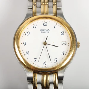 Silver och guld klocka från Seiko - Elegant klocka från Seiko med en kombination av silver och guld i armbandet. Den har en rund urtavla med datumvisning och klassiska visare. Perfekt för en stilren look. Liten repa, därför priset
