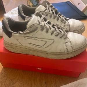 Vita sneakers från Diesel - Säljer ett par vita sneakers från Diesel med snörning. Skorna har en stilren design med svarta detaljer och är perfekta för en avslappnad stil.