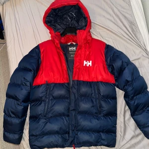 Röd och marinblå pufferjacka från Helly Hansen - Snygg pufferjacka från Helly Hansen i röd och marinblå färg. Jackan har en huva och dragkedja framtill. Perfekt för kyliga dagar.