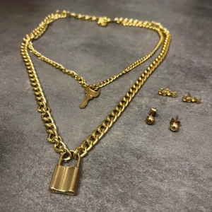 Guldhalsband med lås och nyckel - Stilrent guldhalsband med två kedjor, en med ett hängande lås och den andra med en nyckel. Perfekt för att ge en edgy touch till din outfit. Inkluderar matchande örhängen för en komplett look.