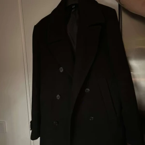Svart kappa/rock från H&M - Elegant svart kappa/rock från H&M med dubbelknäppning och klassisk krage. Lite längre rock i storlek 50 som motsvarar L
