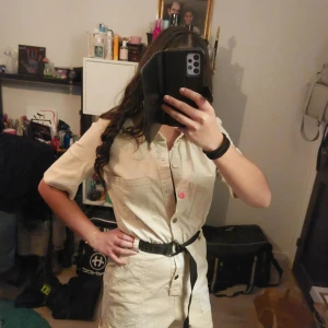 Beige jumpsuit med bälte - Snygg beige jumpsuit med korta ärmar och knappar framtill. Den har ett svart bälte i midjan för en markerad siluett. Perfekt för en stilren look.
