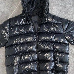 Svart pufferjacka från Livergy - Snygg svart pufferjacka från Livergy med glansig yta och dragkedja framtill. Jackan har en huva och är perfekt för kyligare dagar. Den är designad för att hålla dig varm och bekväm.