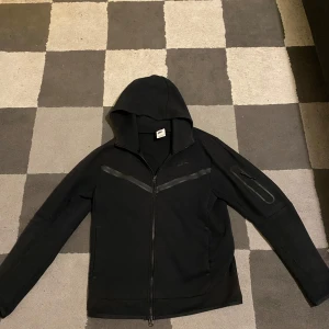Svart Nike tech  - Säljer en svart nike tech med dragkedja uppe och nere och huva. Jackan har långa ärmar och en diskret logga på tröjan, som är ifrån JD Sport.Den har tappat lite färg men annars är den i bra skick. Säljer den då den inte kommer till nån användning längre! Priset kan diskuteras.