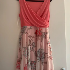 Rosa blommig klänning från Bodyflirt - Charmig rosa klänning från Bodyflirt med omlott-design och blommigt mönster. Klänningen har en v-ringad topp och är ärmlös, vilket ger en luftig känsla. Perfekt för sommarens festligheter.