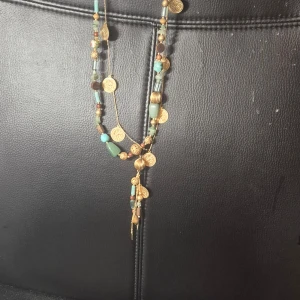 Färgglatt halsband med gulddetaljer - Unikt halsband från DanishDesign med färgglada pärlor och guldfärgade myntdetaljer. Halsbandet har en mix av turkosa, bruna och guldiga pärlor samt dekorativa hängen. Perfekt för att ge en bohemisk touch till din stil.