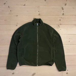 J Lindeberg fleece - Snygg grön J.Lindeberg tröja med dragkedja framtill. Jackan har en hög krage och långa ärmar, perfekt för afterski Den har en stilren design med två fickor på sidorna. Tvättad en gång. Storlek L, men fungerar även för personer som har M.