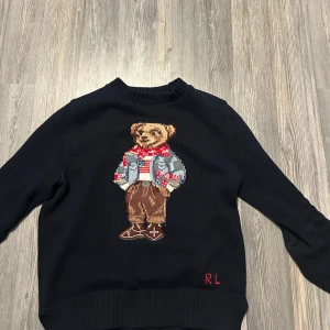 polo bear - Säljer en marinblå stickad tröja från Ralph Lauren med ett coolt nallemotiv på framsidan. Tröjan har långa ärmar och en klassisk rund halsringning. Perfekt för att ge din outfit en unik touch!