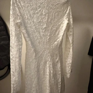 Vit spetsklänning - Elegant vit spetsklänning med långärmad design och v-ringad hals. Perfekt för en stilren och klassisk look. Klänningen har en figurnära passform och är tillverkad i ett vackert spetsmaterial. Passar perfekt till studenten och mottagning.