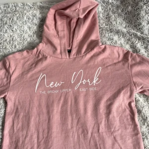 Rosa hoodie från Reaction - Säljer en rosa hoodie från Reaction med texten 'New York' och 'The Bronx Upper East Side' tryckt på framsidan. Den har en mysig huva och är perfekt för en avslappnad stil. Passar bra till både jeans och mjukisbyxor. ALDRIG ANVÄND!