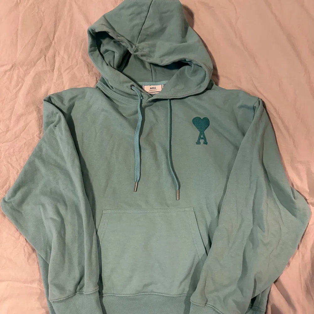 Säljer en snygg ljusblå hoodie från Ami med en stilren design. Hoodien har en känguruficka och en liten hjärtdetalj på bröstet. Perfekt för en avslappnad stil.. Neuletakit & Villapaidat.
