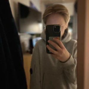 Grå hoodie med ficka - Säljer en stilren grå hoodie med en stor ficka framtill och en luva. Perfekt för en avslappnad look. Den har långa ärmar och en diskret patch på ena ärmen.