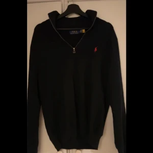 Svart tröja med dragkedja från Polo Ralph Lauren - Snygg svart tröja från Polo Ralph Lauren med dragkedja och röd logga på bröstet. Perfekt för en stilren look. Tröjan har långa ärmar och en klassisk passform.