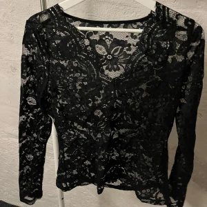 Svart spetsblus - Säljer en elegant svart spetsblus med långärmad design. Blusen har ett vackert blommönster och är perfekt för en stilfull look. Den är tillverkad i ett genomskinligt spetsmaterial som ger en sofistikerad touch.