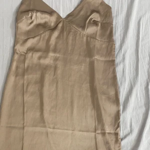 Champagnefärgad klänning från Bubbleroom - Elegant champagnefärgad klänning från Bubbleroom i ett glansigt material. Klänningen har en v-ringad design och är ärmlös, vilket ger en stilren look. Perfekt för speciella tillfällen.