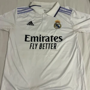  Real Madrid fotbollströja  - Säljer en vit Real Madrid fotbollströja från Adidas med korta ärmar. Tröjan har klubbens emblem och sponsorlogga på framsidan. Storlek S. 