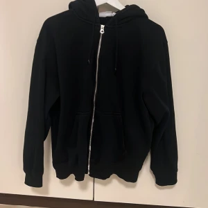 Svart hoodie från Weekday - Säljer en stilren svart hoodie från Weekday i storlek M. Den har en praktisk dragkedja framtill och en bekväm huva med snörning. 