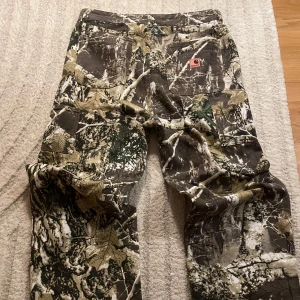 Carhartt Camouflage Jeans - Tja! 😁Säljer ett par  camo carpenter jeans från Carhartt. Byxorna har flera praktiska fickor och en avslappnad passform, perfekt för utomhusaktiviteter🫡… Enda saken dåligt med byxorna är att stäng knappen ovanför Gylfen är lite sne men annars går de fortfarande att stänga utan problem 🫶🏼😁